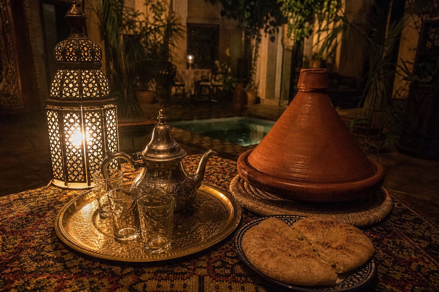 An evening table at Le Vrai Traditionnel, engraved brass tea tray with a silver berrad, three glasses, a sealed terracotta tagine on a palm-fibre mat, fresh khobz on a small plate.