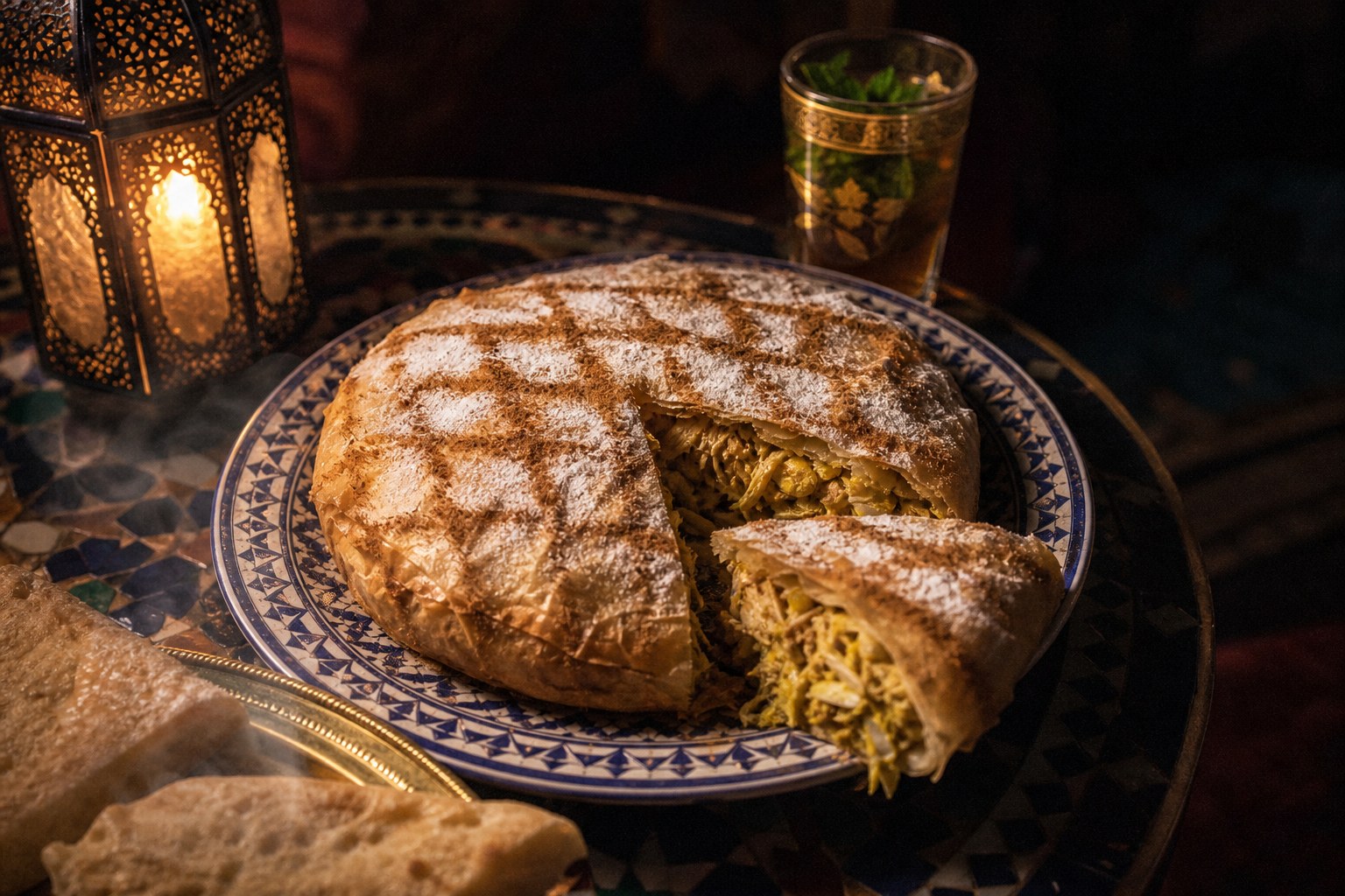 A Moroccan pastilla on a zellige plate, golden warqa dusted with powdered sugar and cinnamon, sliced to reveal the spiced filling.
